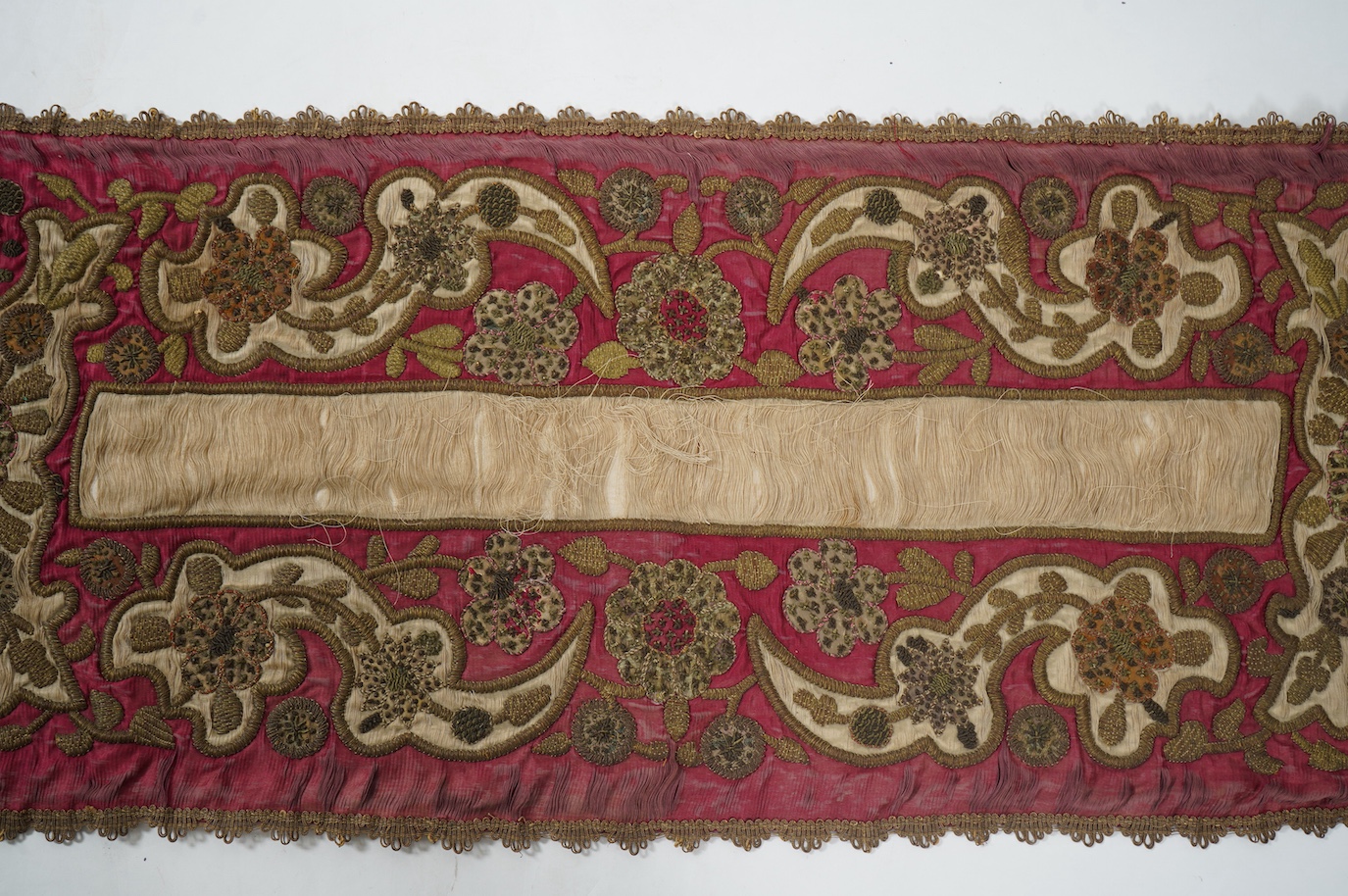 A 19th century Indian red and cream silk runner, heavily embroidered with scrolling motifs, using a variety of stump work and gold wire work stitches, with earlier Mughal influences, 118cm x 46.5cm (not including braid),